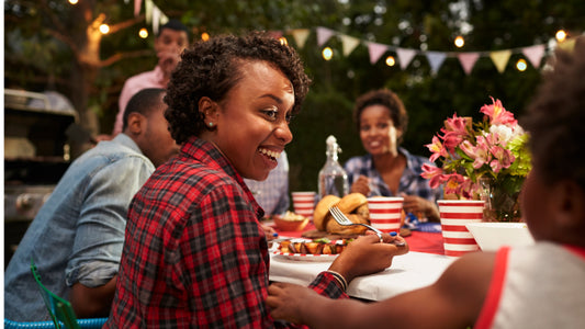 A family enjoys a 4th of July celebration meal together at an outdoor table with lights and streamers in the background