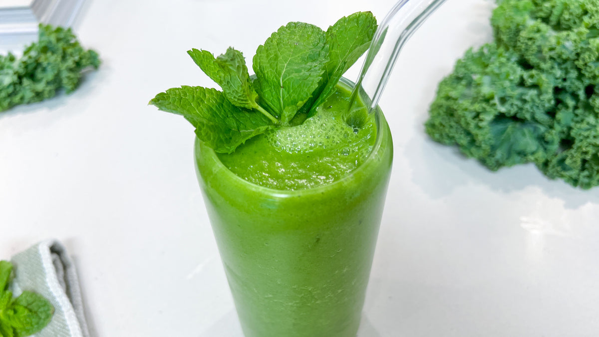 A green smoothie in a glass with glass straw surrounded by kale and mint leaves 