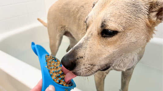 A tan dog in a bath tub eats peanut butter from a lick mat