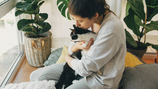 A woman snuggling with a cat while sitting on pillows in front of plants