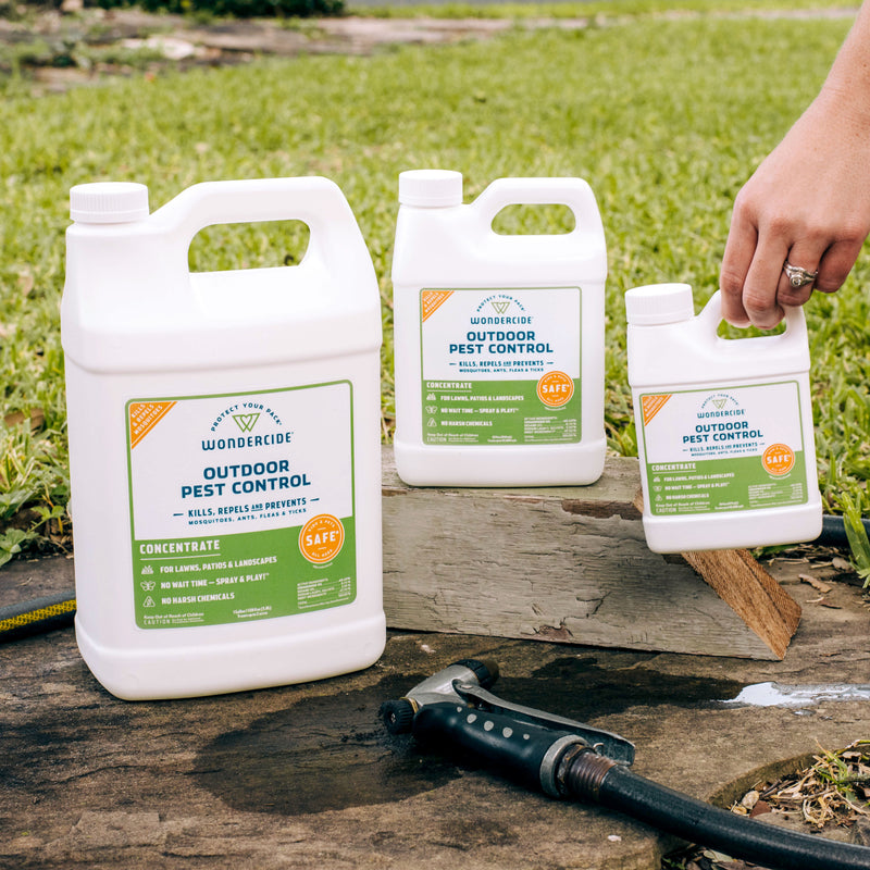 A 128 oz bottle of Wondercide Outdoor Pest Control Concentrate for Yard + Garden sits on a stone surface outside, ready to help you tackle mosquitoes and pests with ease. Grass and a garden hose are nearby, making application simple.