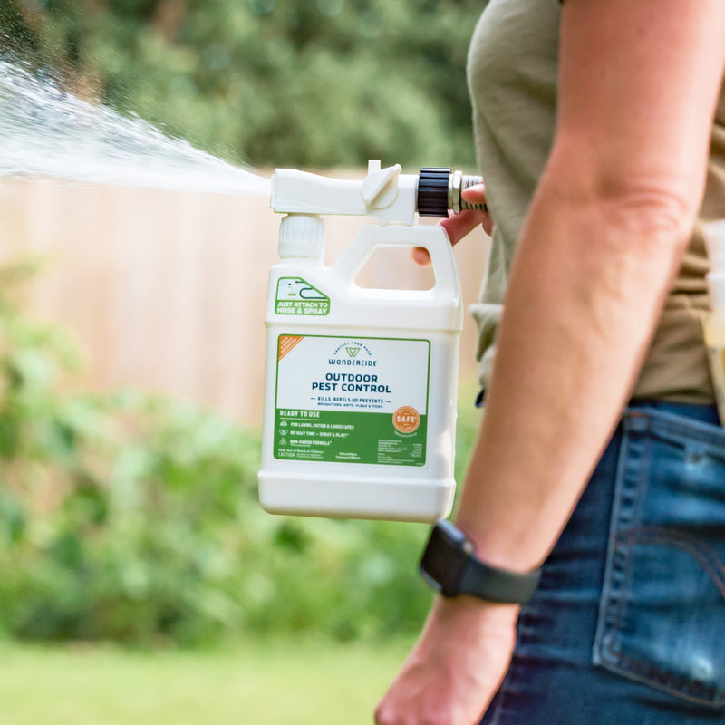 A person in a tan shirt and blue jeans sprays Wondercide Ready-to-Use Outdoor Mosquito Spray from a white bottle with hose attachment in the backyard, effectively controlling pests like fleas and mosquitoes. Visible spray can be seen coming from the nozzle.
