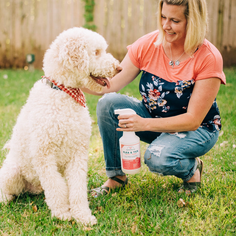 A woman kneels on grass beside a large white dog in a red checkered bandana, smiling as she holds Wondercide Peppermint Flea & Tick Spray for Pets + Home, labeled safe for dogs and cats, outdoors near a wooden fence.