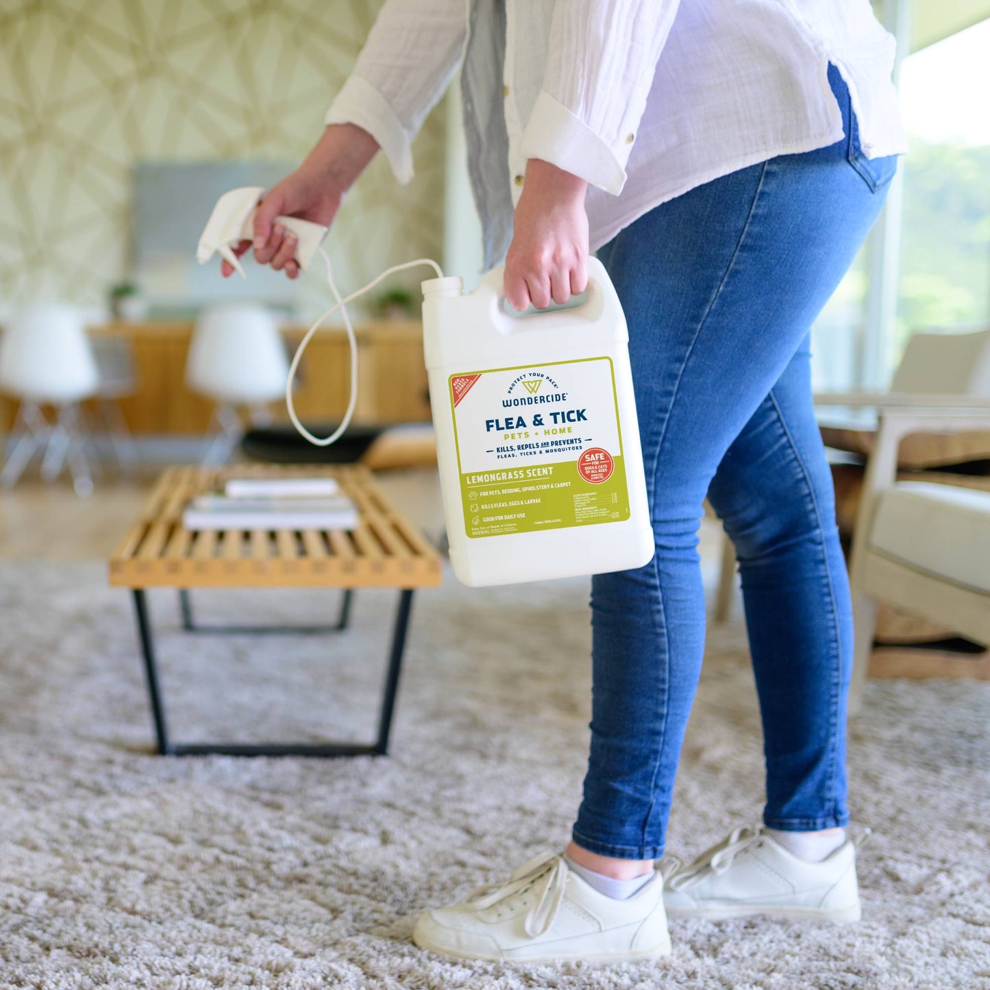 A person in jeans and white sneakers holds a 128 oz. Wondercide Lemongrass Flea & Tick Spray for Pets + Home with Natural Essential Oils while standing on a carpeted floor in a modern living room.