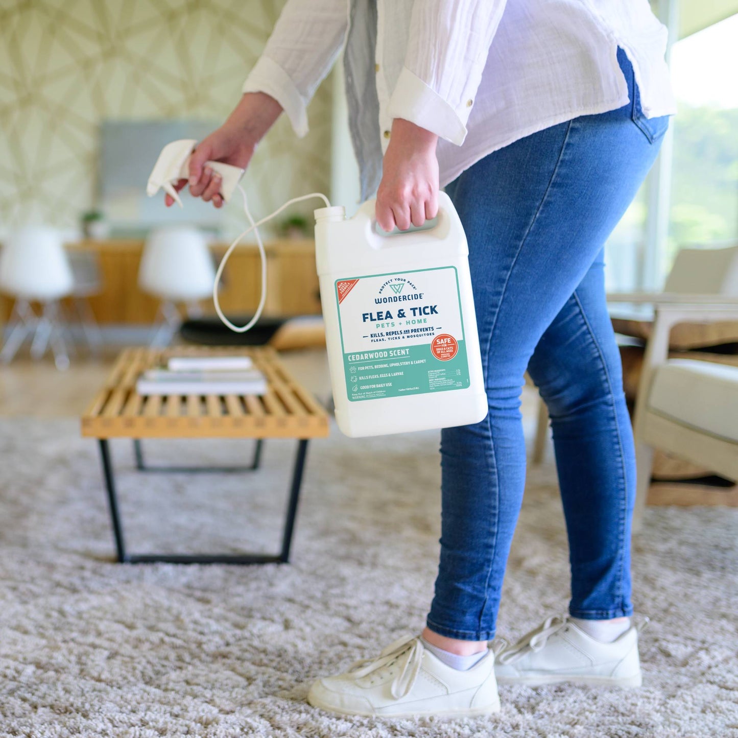 A person in jeans and white sneakers holds a large 128 oz. Wondercide Cedarwood Flea & Tick Spray for Pets + Home with natural essential oils, standing in a bright, modern living room with a rug and wooden coffee table.