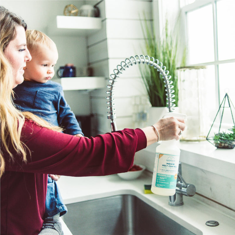 A woman uses Wondercide 128 oz. Cedarwood Indoor Pest Control, safe around pets, as she sprays her kitchen sink by a sunny window with plants, holding her toddler and enjoying the fresh cedarwood scent.