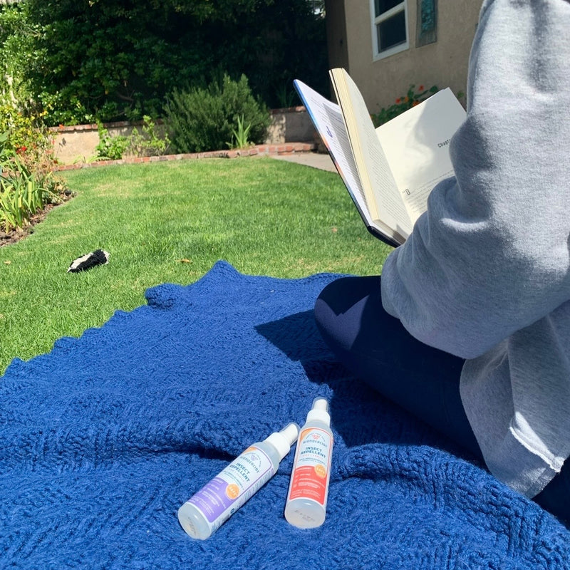 A person sitting on a blue blanket in a grassy yard is reading a book. Two bottles of sunscreen are on the blanket. There are bushes and a small building in the background under a clear sky.