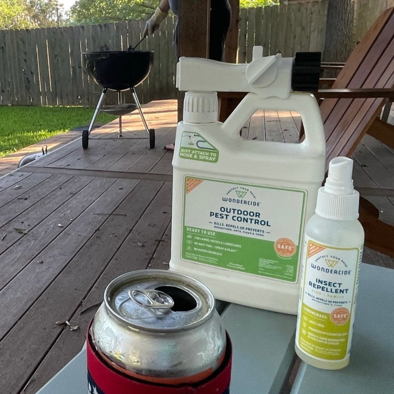 Outdoor scene with a wooden deck, a can of soda in a koozie, and two pest control products on a table. A barbecue grill is visible in the background near a wooden fence and trees.