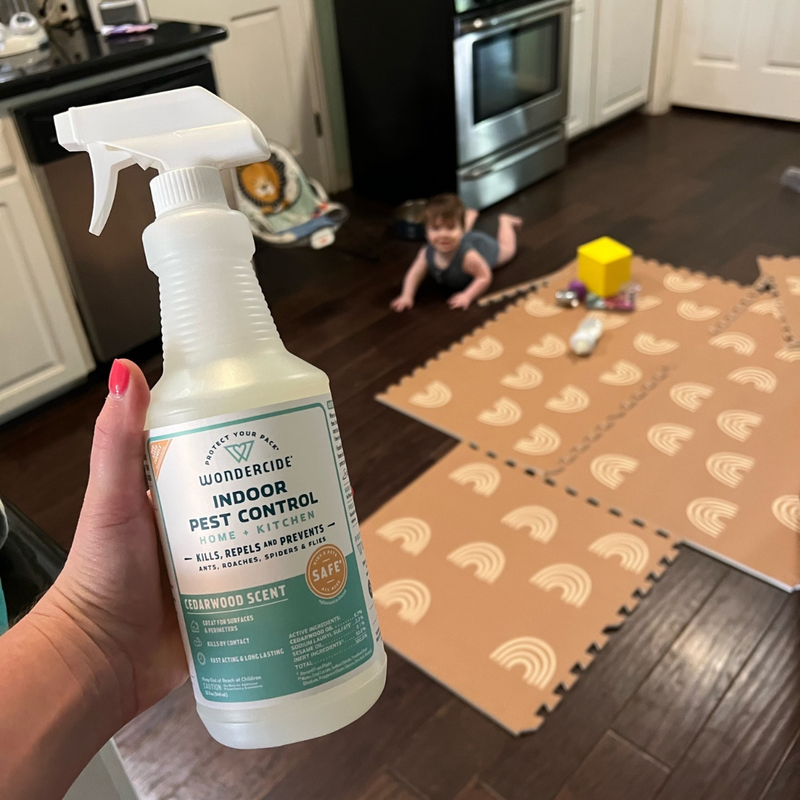 A person holds a spray bottle labeled Wondercide Indoor Pest Control in a kitchen. A baby crawls on patterned foam mats nearby, with toys scattered around. The setting suggests a home environment.