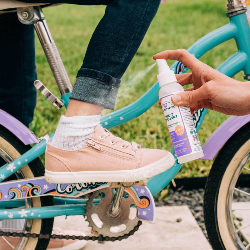 An adult sprays Wondercide Rosemary Insect Repellent for Family on a child's ankle as the child, wearing pink shoes and rolled-up jeans, sits on a blue and purple bike, enjoying protective outdoor fun in grassy surroundings.