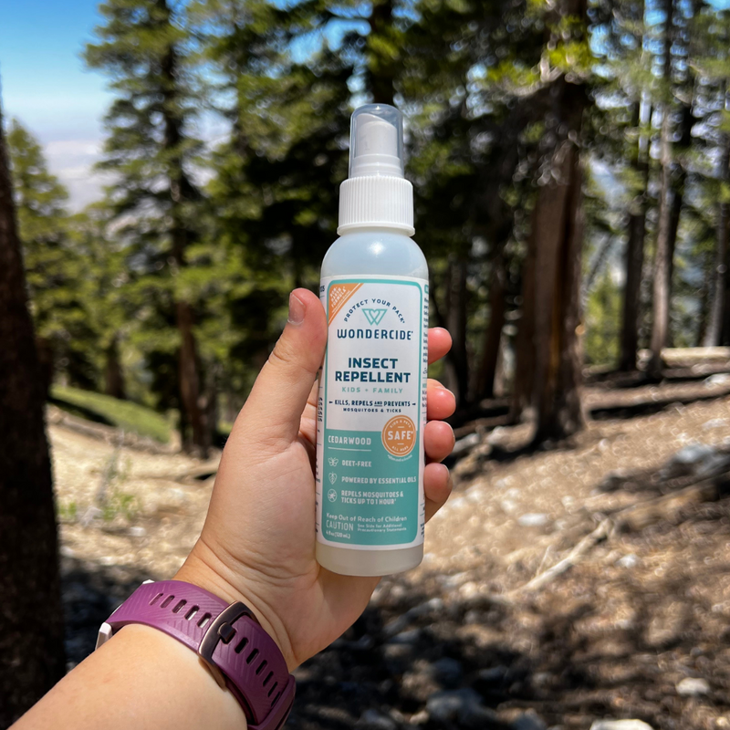 A hand holds a bottle of Wondercide insect repellent in a forest setting. The product label highlights its natural cedarwood scent and DEET-free formula. Tall trees and a glimpse of the sky are in the background. A purple wristband is visible.