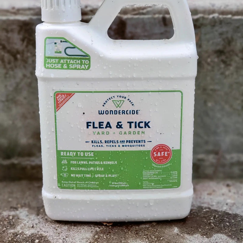 A container of Wondercide Flea & Tick spray placed on a concrete surface. The label indicates it is for yard and garden use and claims to kill, repel, and prevent fleas, ticks, and mosquitoes. The container is covered in small water droplets.