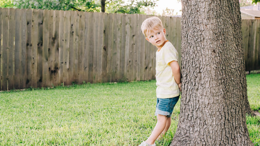 A boy in jean shorts and a yellow shirt plays hide and seek in his backyard