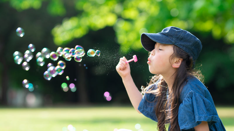 A little girl in a blue denim short sleeve shirt and cap blows irridescent bubbles into the air