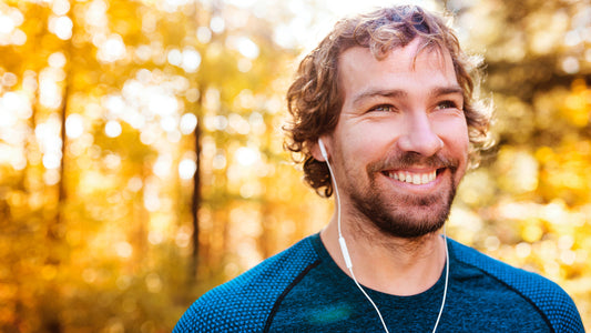 A man in a blue shirt with curly brown hair listens to headphones in the fall outside with trees in the background