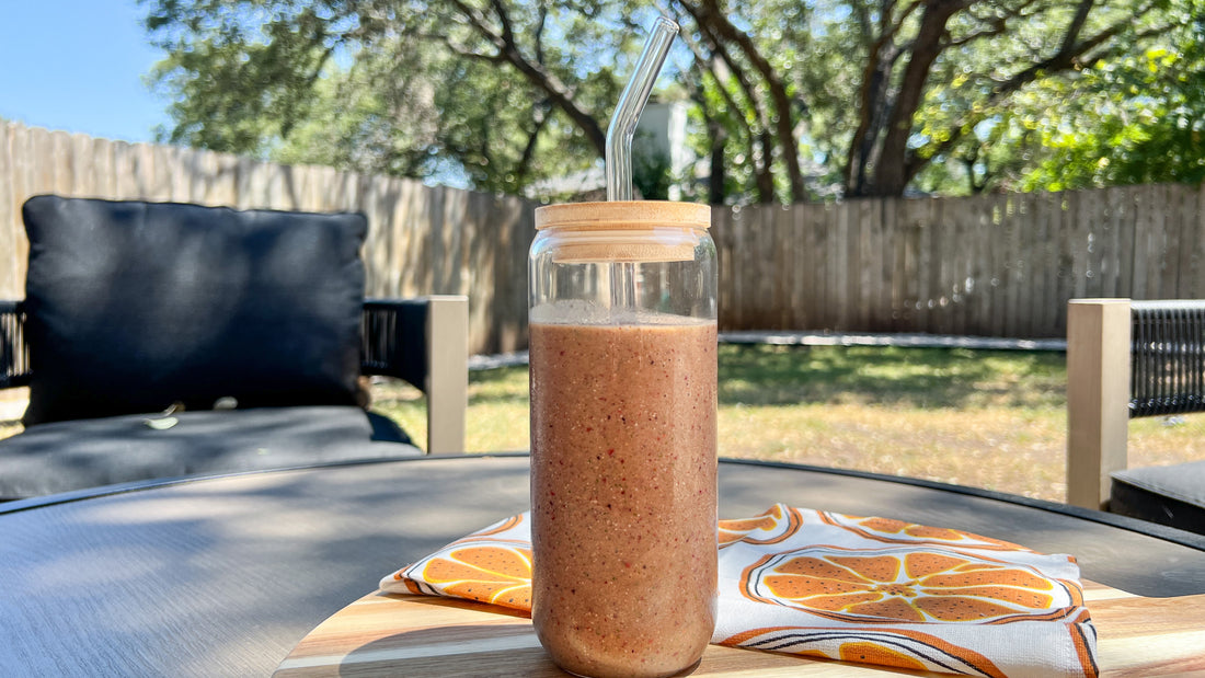 A smoothie in a glass bottle with a glass straw sits on top of a table outside with a tangerine towel next to it