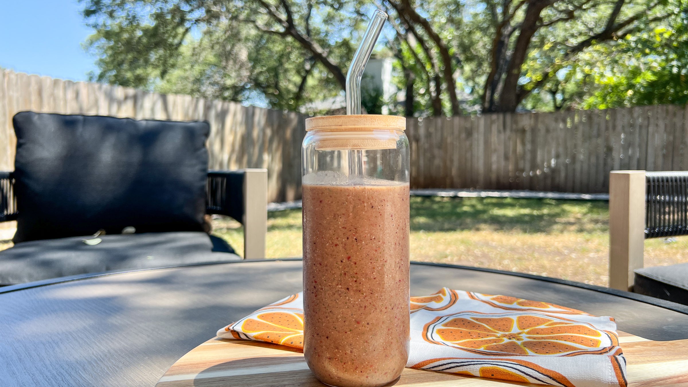 A smoothie in a glass bottle with a glass straw sits on top of a table outside with a tangerine towel next to it