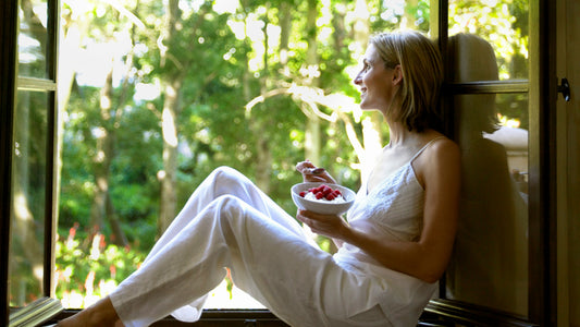 A woman in white pahamas sitting in an open window eating raspberries and yogurt out of a white bowl