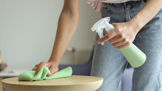A woman wearing jeans wipes the top of a light oak table using a green microfiber cloth and green spray bottle