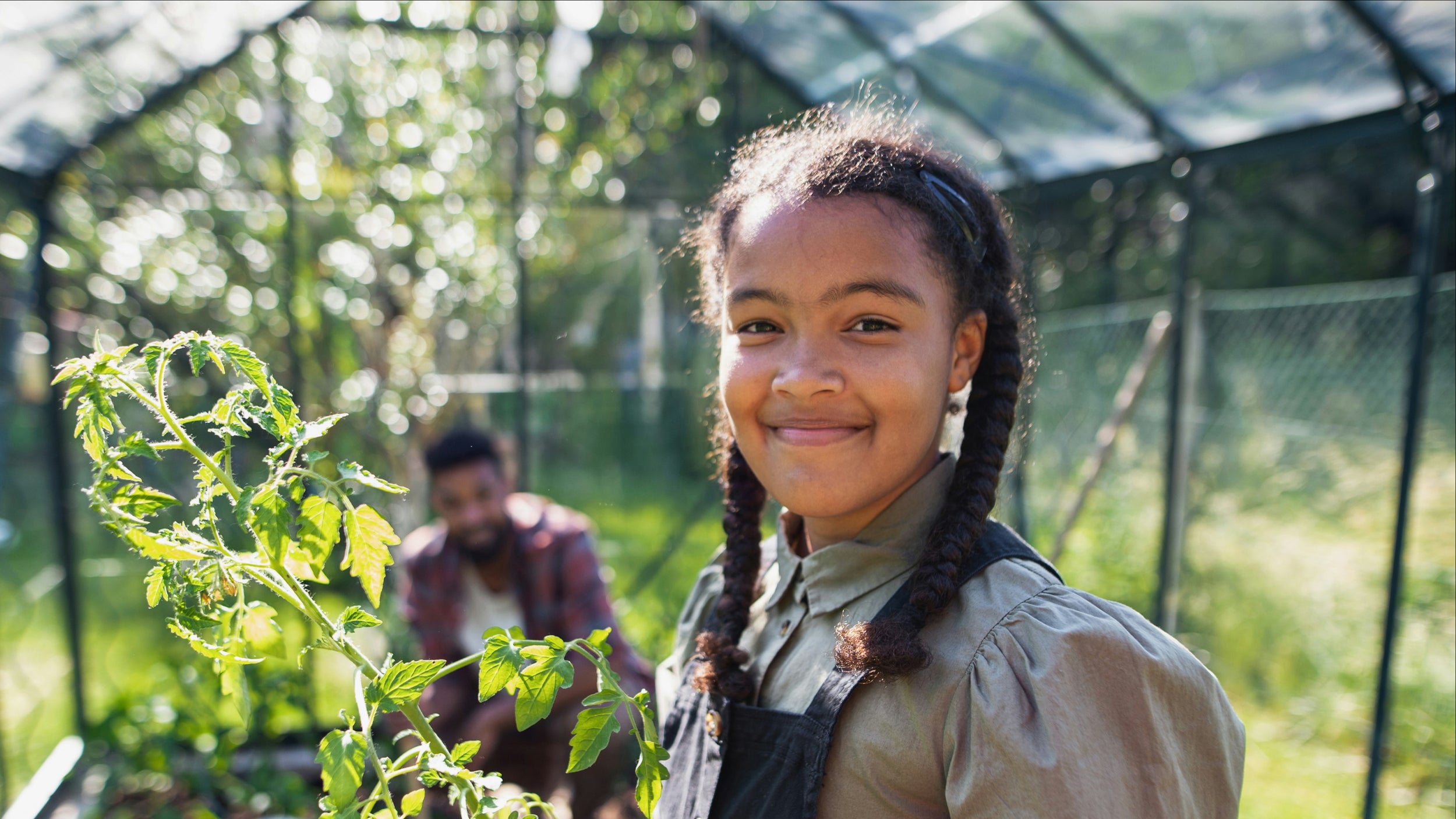 A young girl smiling iwth and pig tails in a greenhouse with a person in the background