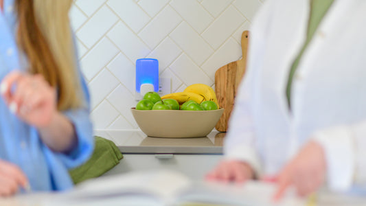 Two people in the foreground look at a cookbook with a bowl of fruit and Wondercide Fruit Fly Trap in the background