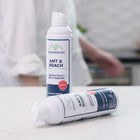 Two cans of Wondercide Ant & Roach for Home + Kitchen sit on a white countertop—one upright, one lying down. A blurred person is in the background. Safe for pets, it kills ants and roaches without harsh chemicals.