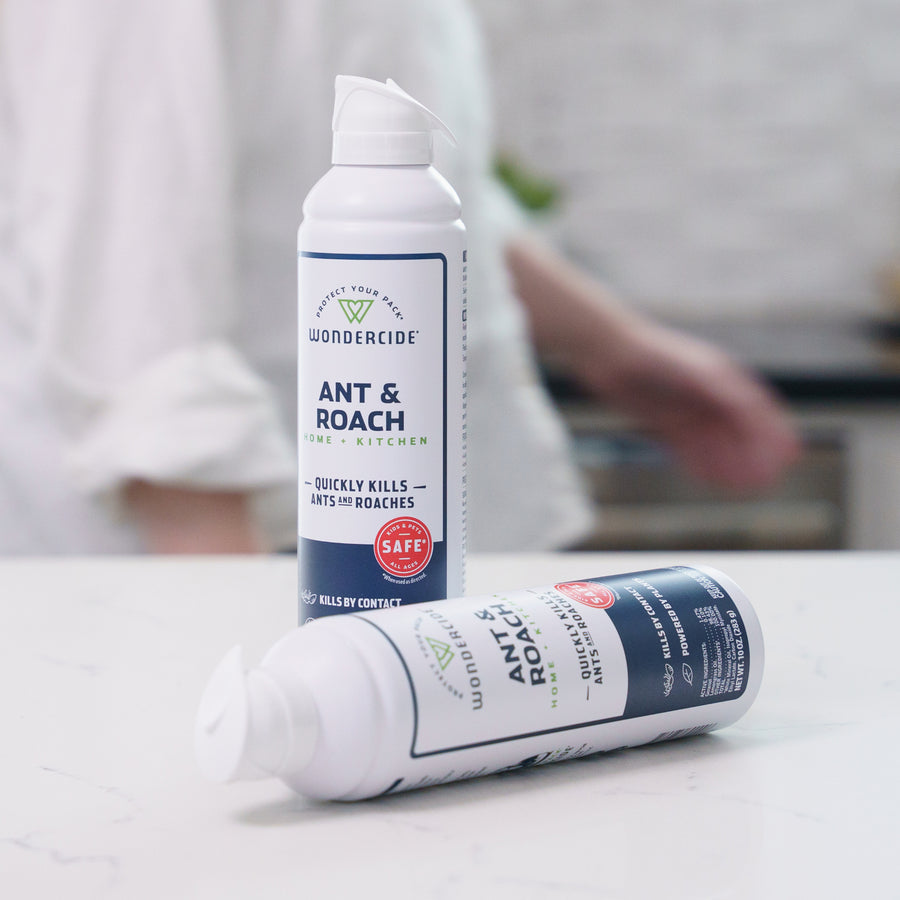 Two cans of Wondercide Ant & Roach for Home + Kitchen sit on a white countertop—one upright, one lying down. A blurred person is in the background. Safe for pets, it kills ants and roaches without harsh chemicals.