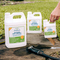 Three containers of Wondercide Mosquito Yard Spray Refill Starter Kit, in various sizes, rest on an outdoor wooden surface with grass behind. A hand holds the smallest bottle and a black hose nozzle is nearby. Plant-powered protection by Wondercide.