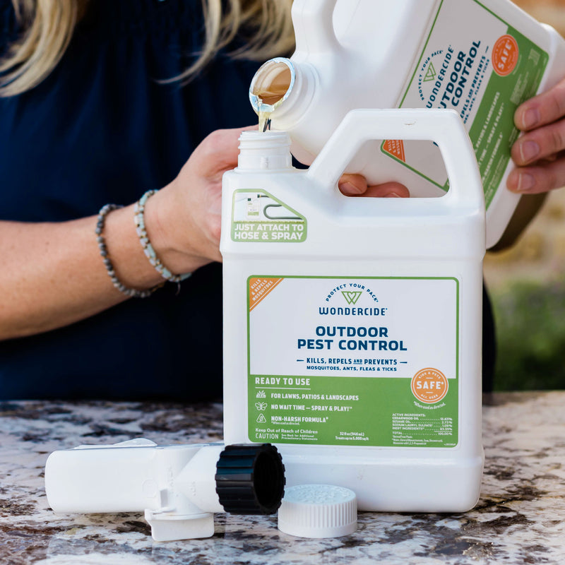 A person pours liquid from a Wondercide Mosquito Yard Spray Refill Starter Kit. A hose attachment and cap are on the table. The container has green and white labeling.