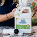 A person pours Wondercide Outdoor Pest Control Concentrate for Yard + Garden, 128 oz, into a bottle on a marble surface to prepare mosquito repellent, with a hose spray nozzle nearby.
