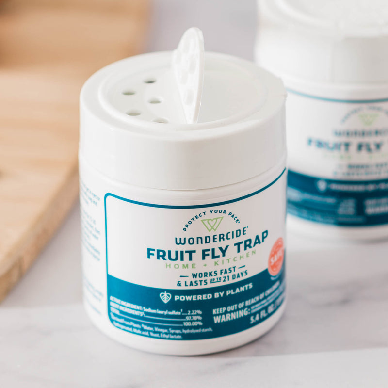 A white container of Wondercide (2 Pack) Fruit Fly Trap for Home + Kitchen with a blue and green label sits on a counter with its open lid showing holes for catching fruit flies. A second, similar container is blurred in the background.