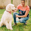 A woman kneels on grass beside her large white curly-haired dog in a red bandana, holding Wondercide Cedarwood Flea & Tick Spray for Pets + Home, in a sunny backyard with a wooden fence behind them.