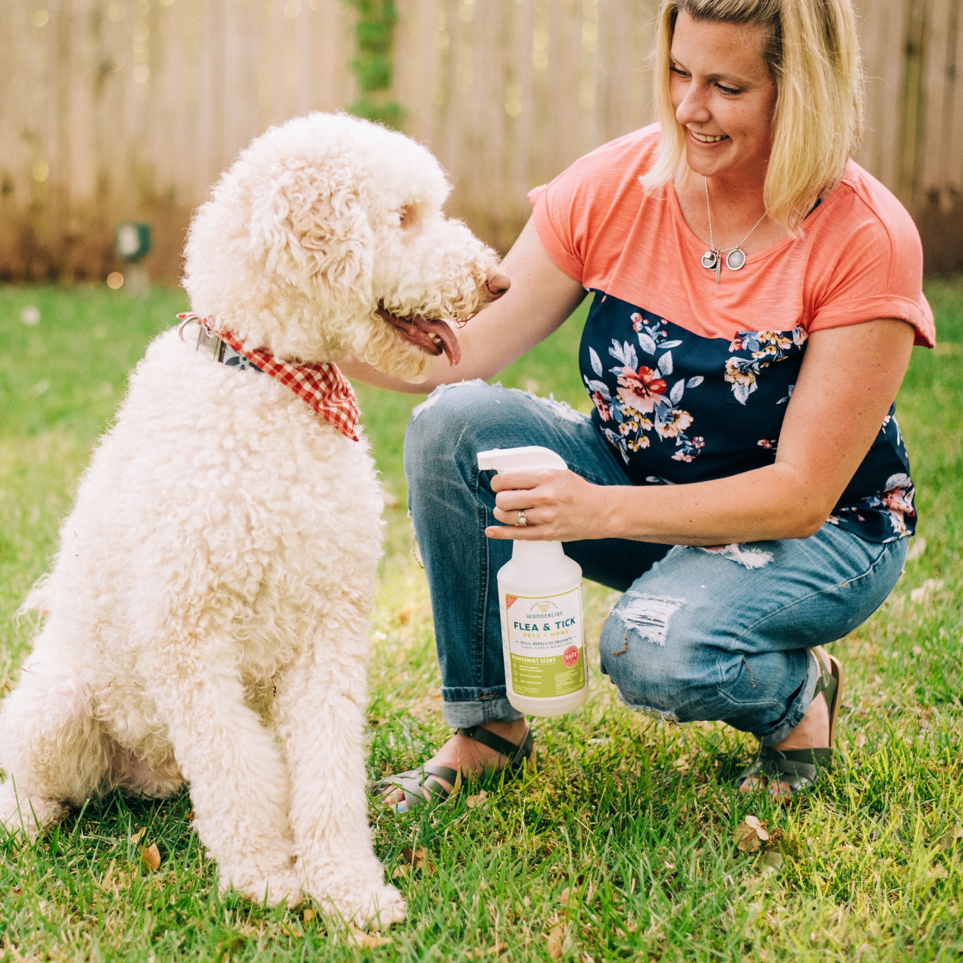 A woman kneels on the grass, smiling at a large, curly-haired dog wearing a red checkered bandana. She holds a Wondercide Pet Parent Pack | Shark Tank Bundle with Natural Essential Oils spray bottle. A wooden fence and greenery enhance their joyful moment.