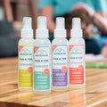 Four bottles of Wondercide Cedarwood Flea & Tick Spray for Pets + Home are arranged in a row on a wooden surface, with blurred people and a dog in the background.
