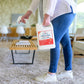 A person in jeans and white sneakers holds a large 128 oz. Wondercide Peppermint Flea & Tick Spray for Pets + Home with Natural Essential Oils, standing on a carpeted floor in a bright, modern living room.