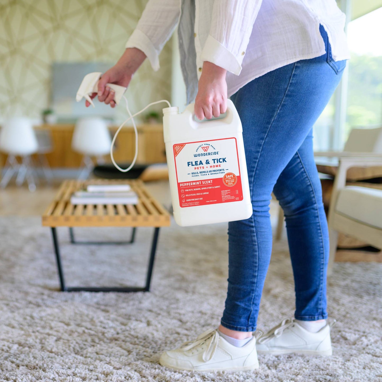 A person in jeans and white sneakers holds a large 128 oz. Wondercide Peppermint Flea & Tick Spray for Pets + Home with Natural Essential Oils, standing on a carpeted floor in a bright, modern living room.