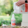 A hand pours Wondercide 128 oz. Peppermint Flea & Tick Spray, made with natural essential oils and pet-safe, from a large jug into a smaller spray bottle on a wooden surface with a green outdoor background.