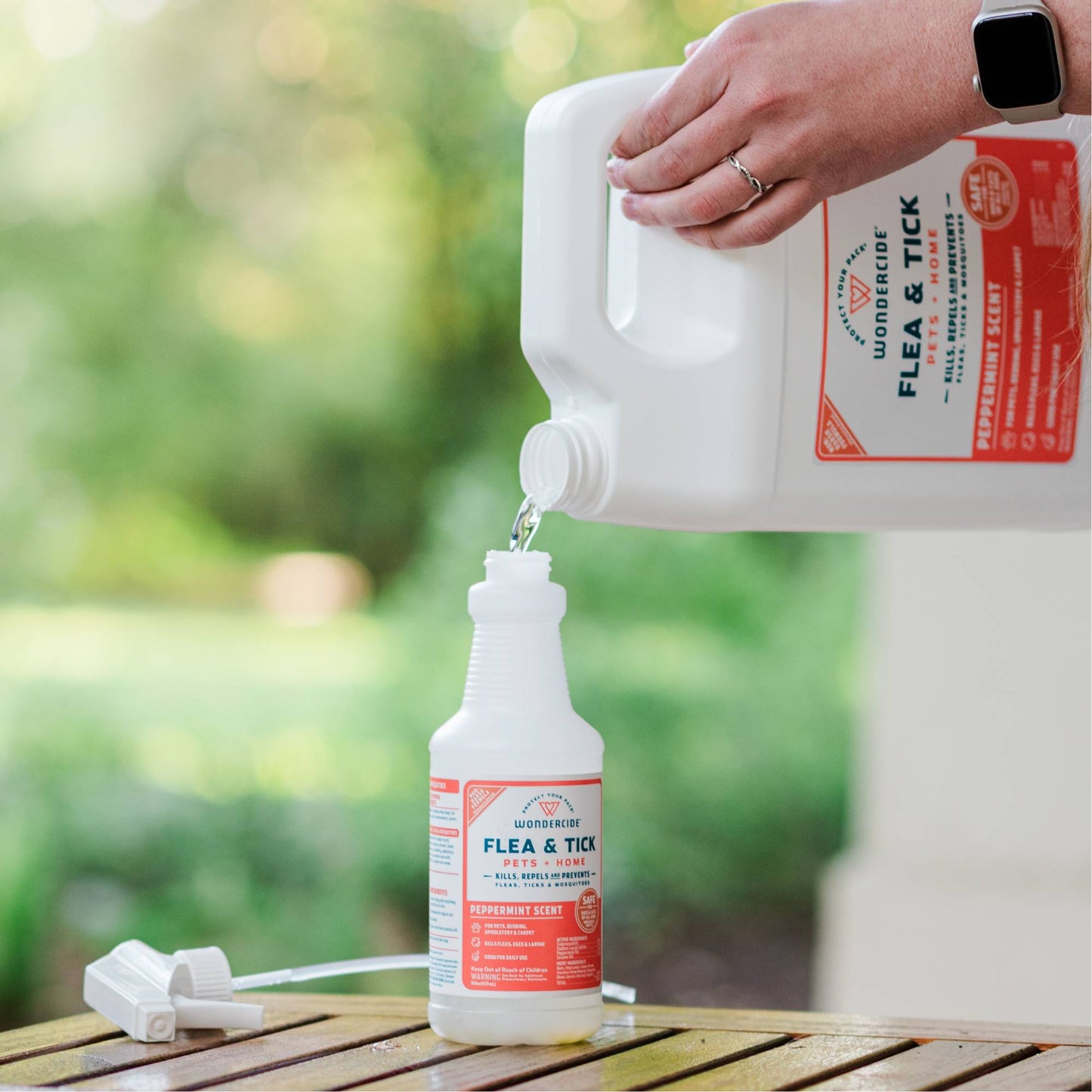 Someone pours Wondercide 128 oz. Peppermint Flea & Tick Spray for Pets + Home with Natural Essential Oils into a smaller spray bottle on a wooden surface, with a green outdoor background.