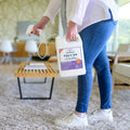 A person in jeans and white sneakers holds a 128 oz. Wondercide Rosemary Flea & Tick Spray made with natural essential oils, preparing to treat the carpeted floor in a bright living room. Safe for pets and tough on fleas and ticks.