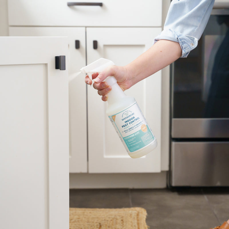 A person sprays Wondercide Indoor Pest Control for Home + Kitchen onto a white kitchen cabinet door in a kitchen with white cabinets, a stainless steel oven, and a woven rug. The spray is pet-safe.