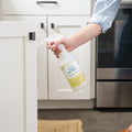 A person holding Wondercide Lemongrass Indoor Pest Control for Home + Kitchen, about to clean a white cabinet door in a kitchen with white cabinets and dark flooring; safe around pets for worry-free use.