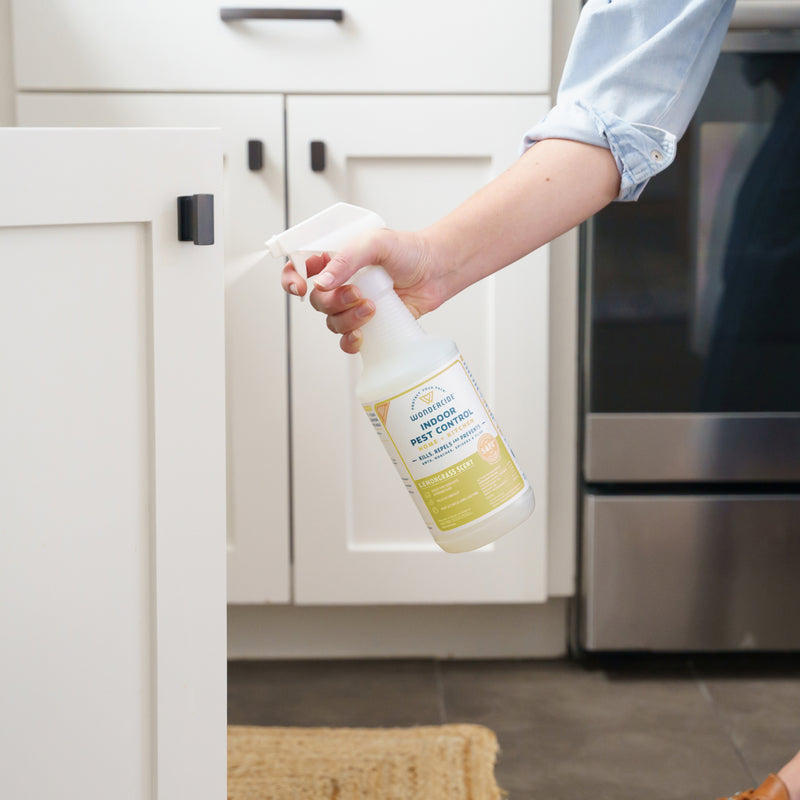 A person holding Wondercide Lemongrass Indoor Pest Control for Home + Kitchen, about to clean a white cabinet door in a kitchen with white cabinets and dark flooring; safe around pets for worry-free use.
