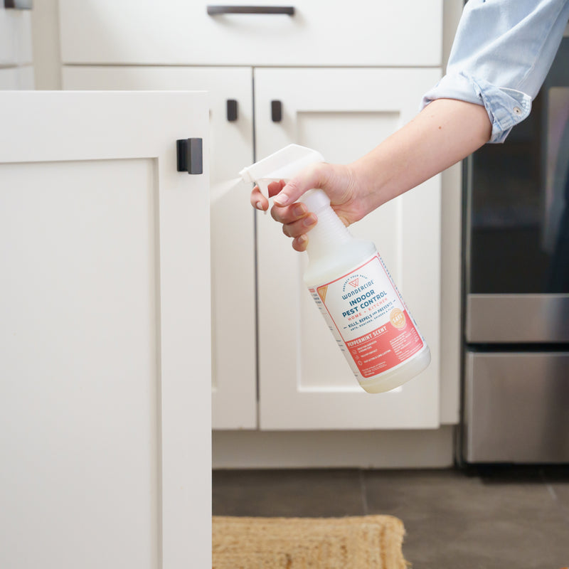 A person sprays Wondercide Peppermint Indoor Pest Control for Home + Kitchen on a white cabinet in a kitchen with dark tile floors and white cabinetry, providing pet-safe indoor pest protection.