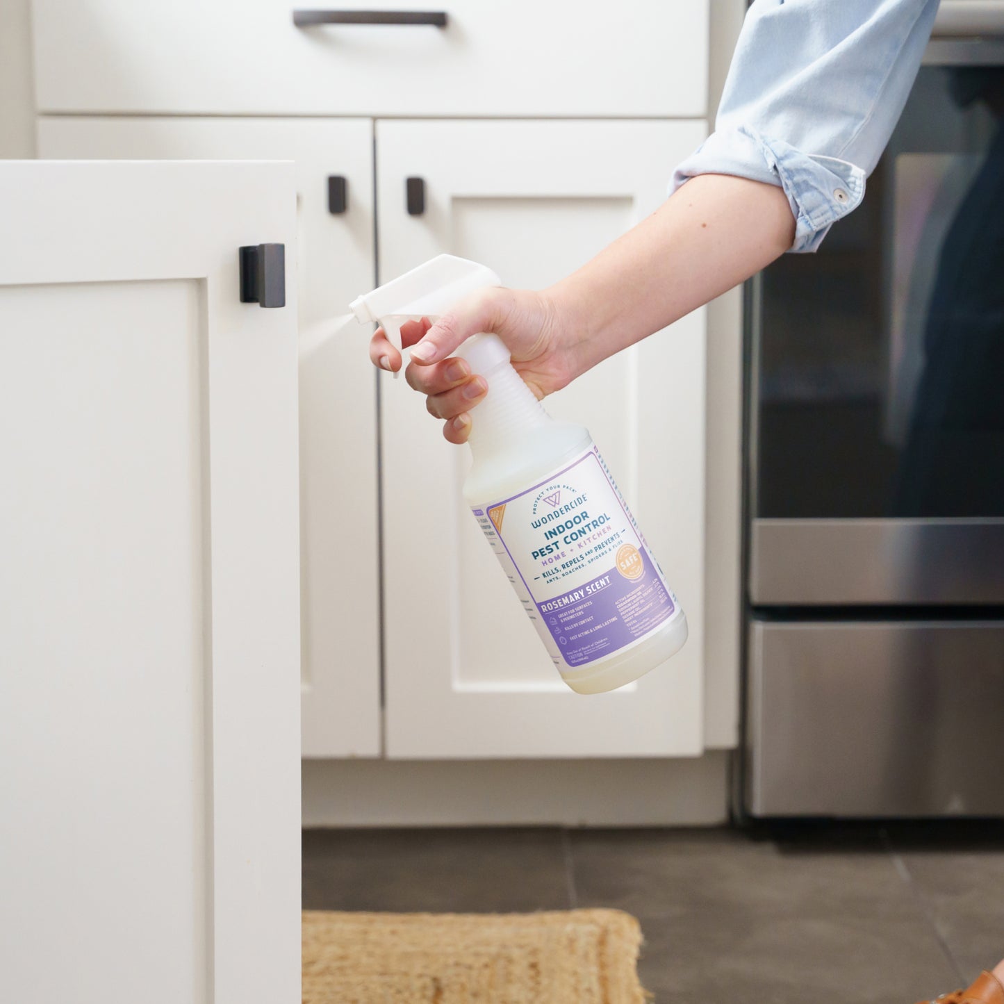 A person in a blue shirt uses Wondercides Pest Protection Kit with Natural Essential Oils to spray pet odor remover onto a white kitchen cabinet. The bottle has a purple label, and the background shows a stainless steel appliance and a woven mat on the floor.
