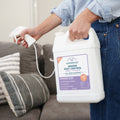 A person holds a large white spray bottle of Wondercide Rosemary Indoor Pest Control for Home + Kitchen, with a gray couch and striped pillows in the living room background.