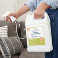 A person holds a large Wondercide Lemongrass Indoor Pest Control for Home + Kitchen jug with a spray nozzle, standing in a living room by a beige couch. The label notes it's safe around pets and perfect for use throughout the home.