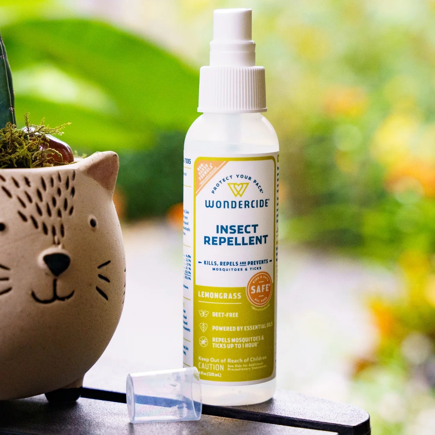 A bottle of Wondercide Lemongrass Insect Repellent for Family with Natural Essential Oils and a white spray cap sits beside a cat-shaped planter, its label emphasizing the fresh lemongrass scent amid softly blurred greenery.