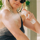 A woman in sunglasses and a black tank top sprays Wondercide Insect Repellent for Family on her arm outdoors, surrounded by sunlight and greenery.