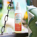 A bottle of Wondercide Peppermint Insect Repellent for Family rests on a wooden surface beside a green and white pillow, with a metal chain and blurred greenery in the background.
