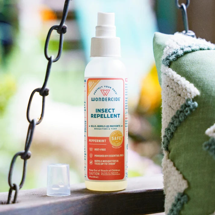 A bottle of Wondercide Peppermint Insect Repellent for Family rests on a wooden surface beside a green and white pillow, with a metal chain and blurred greenery in the background.